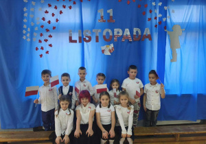 11 dzieci stoi na tle niebieskiej dekoracji. W dłoniach trzymają flagi.