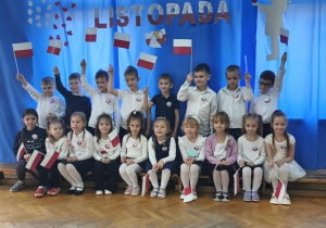 Dzieci stoją i siedzą na tle niebieskiej dekoracji. Część dzieci trzyma w dłoniach flagi.