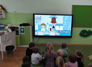 Dzieci siedzą na zielonym dywanie i oglądają film edukacyjny.