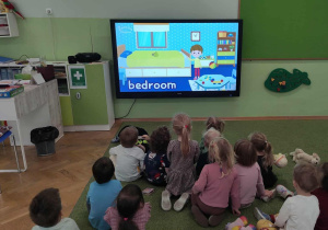 Dzieci siedzą na zielonym dywanie i oglądają film edukacyjny.