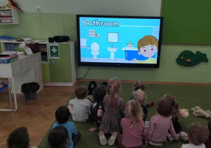 Dzieci siedzą na zielonym dywanie i oglądają film edukacyjny.