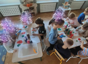 Dzieci siedzą przy stolikach i malują farbami.