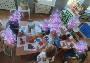 Dzieci siedzą przy stolikach i malują farbami.