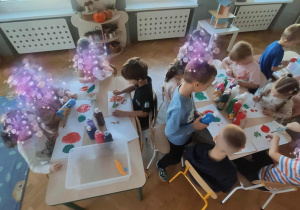 Dzieci siedzą przy stolikach i malują farbami.