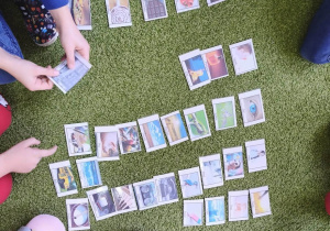 Na zielonym dywanie leżą kolorowe karty. Wokół widać nogi siedzących dzieci.