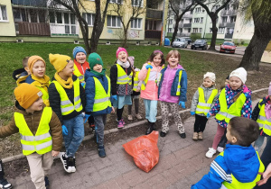 Grupa dzieci w kamizelkach stoi na chodniku. W środku stoi czerwony worek ze śmieciami.