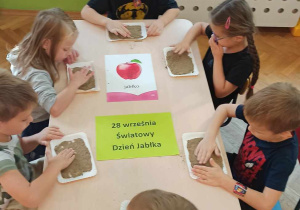 Dzieci siedzą przy stoliku i rysują na piasku kształt jabłka