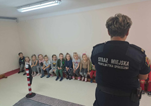 Dzieci siedzą na ławkach i słuchają przedstawiecieli Straży Miejskiej