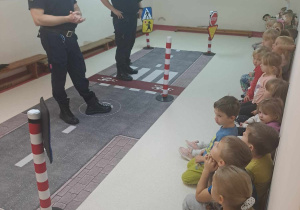 Dzieci siedzą na ławkach i słuchają przedstawiecieli Straży Miejskiej