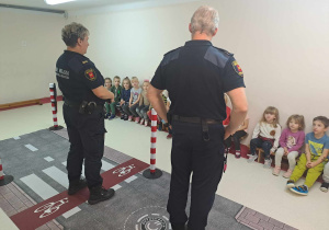 Przedstawiciele Straży Miejskiej stoją i przypominają dzieciom zasady bezpieczeństwa, dzieci siedzą na ławkach
