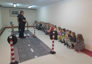 Dzieci siedzą na ławkach i słuchają przedstawieciela Straży Miejskiej