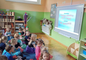 Dzieci siedzą na dywanie przed tablicą multimedialną gdzie wyświetlana jest bajak
