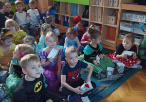 Dzieci siedzą na dywanie i krzesełkach jedzą popcorn i oglądają seans