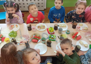 Dzieci siedzą przy stolikach i jedzą samodzielnie przygotowane kanapki