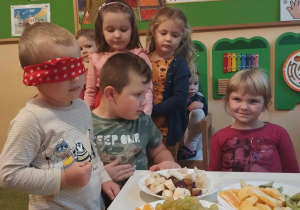 Chłopiec z zawiązanymi oczami stoi przy stoliku z owocami.