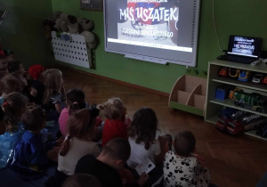 Dzieci siedzą na materacach i oglądają bajkę na tablicy mulimedialnej.