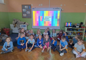 Grupa dzieci siedzi na podłodze i pozuje do zdjecia w tle tablica multimedialna