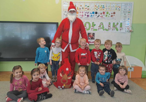 Grupa dzieci pozuje do zdjęcia ze Świętym Mikołajem w tle napis Mikołajki