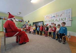 Mikołaj siedzi na krześle, dzieci stoją w szeregu i śpiewają dla niego piosenkę