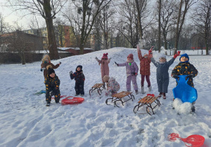Grupa dzieci pozuje do zdjecia w ogrodzie przedszkolnym, przed dziećmi stoją sanki