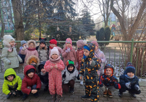 Grupa dzieci pozuje do zdjęcia na tle drzewa na którym wisi karmnik z ziarnami dla ptaków