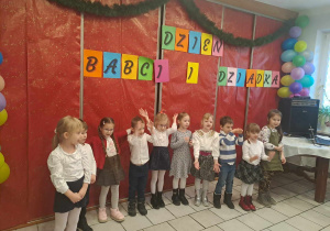 Dzieci ubrane na galowo stoją w szeregu w tle napisz Dzień Babci i Dziadka