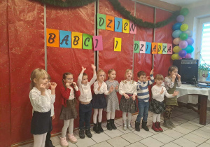 Dzieci ubrane na galowo stoją w szeregu w tle napisz Dzień Babci i Dziadka