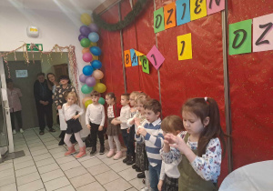 Dzieci ubrane na galowo stoją w szeregu w tle napisz Dzień Babci i Dziadka