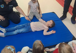 Dziewczynka ćwiczy układanie poszkodowanego w pozycji bezpiecznej, dzieci patrzą.