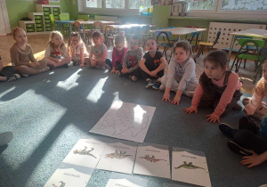 Dzieci siedzą na dywanie i słuchają informacji o dinozaurach