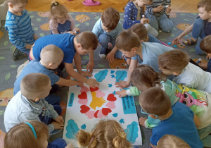 Dzieci siedzą na szarym dywanie i układają niebieskie puzzle na białym bristolu.