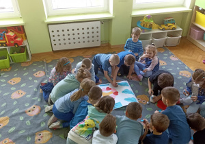 Dzieci siedzą na szarym dywanie i układają niebieskie puzzle na białym bristolu. Dwójka dzieci trzyma w rękach kleje.