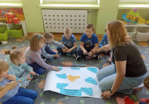Dzieci siedzą na szarym dywanie. W środku leży biały bristol z niebieskimi napisami. Jedna z dziewczynek nakleja napis.