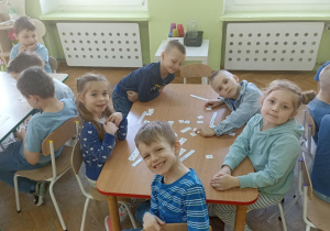 Piątka dzieci siedzi przy stoliku, uśmiecha się do zdjęcia i układa napis z białych kartoników.