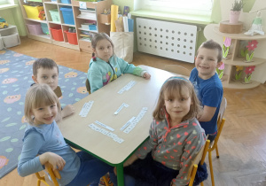 Piątka dzieci siedzi przy stoliku z ułożonymi napisami z białych kartoników. Z lewej strony zdjęcia widać na podłodze szary dywan w sowy.