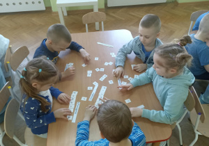 Piątka dzieci siedzi przy stoliku i układa napis z białych kartoników.