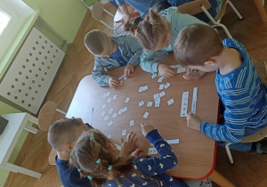 Piątka dzieci siedzi przy stoliku i układa napis z białych kartoników. Z lewej strony zdjęcia widać biały stolik z kolorowymi kubkami i dzbankiem z wodą.