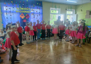 Dzieci w czerwonych koszulkach i różowych spódniczkach stoją w półkolu.