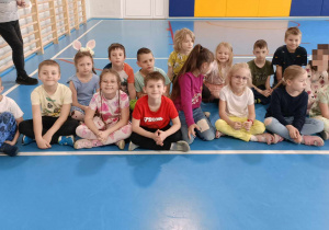Dzieci siedzą na niebieskiej podłodze w sali gimnastycznej. Po lewej stronie widać przymocowane do ściany drabinki.