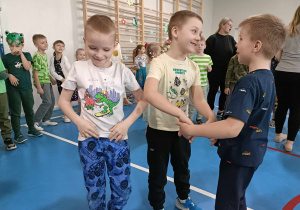Chłopiec w niebieskich spodniach tańczy sam. Dwóch chłopców obok niego tańczy w kółeczku.