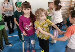 Dziewczynka w fioletowej bluzie uśmiecha się do zdjęcia. Dwóch chłopców tańczy w kółeczku.