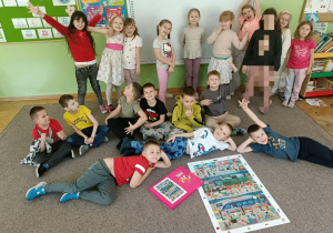 Grupa dzieci stoi i siedzi na dywanie z ułożonymi puzzlami. W tle widać białą tablicę.