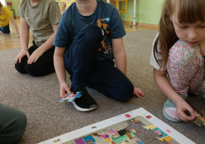 Chłopiec siedzi na dywanie, układa puzzle i uśmiecha się do zdjęcia. Z prawej strony siedzi dziewczynka a z tyłu siedzi chłopiec.