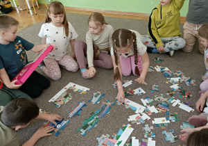 Wokół puzzli siedzi jedenastka dzieci. Chłopiec w granatowej bluzce trzyma w dłoniach różowe pudełko.