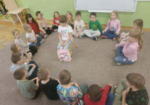 Dzieci siedzą w kole. W środku koła na żółtym krzesełku siedzi dziewczynka z różową torbą.