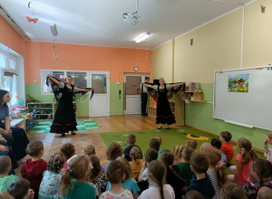 Dwie panie tańczą w czarnych sukienkach z czarnymi chustami w dłoniach. Na dywanie siedzą dzieci. Po prawej stronie widać białą tablicę.