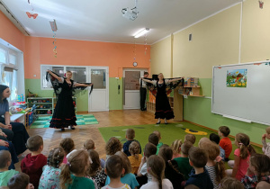 Dwie panie tańczą w czarnych sukienkach z czarnymi chustami w dłoniach. Na dywanie siedzą dzieci. Po prawej stronie widać białą tablicę.