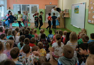 Dzieci tańczą w kole z baletnicami. Na widowni siedzą dzieci. Po prawej stronie widać białą tablicę.