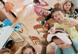 Dzieci siedzą na krzesełkach i machają do zdjęcia. W tle widać stolik.