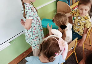 Dziewczynka w niebieskiej koszulce stoi na dywanie z niebieskim pudełkiem. Na krzesełkach siedzą dwie dziewczynki oraz chłopiec.